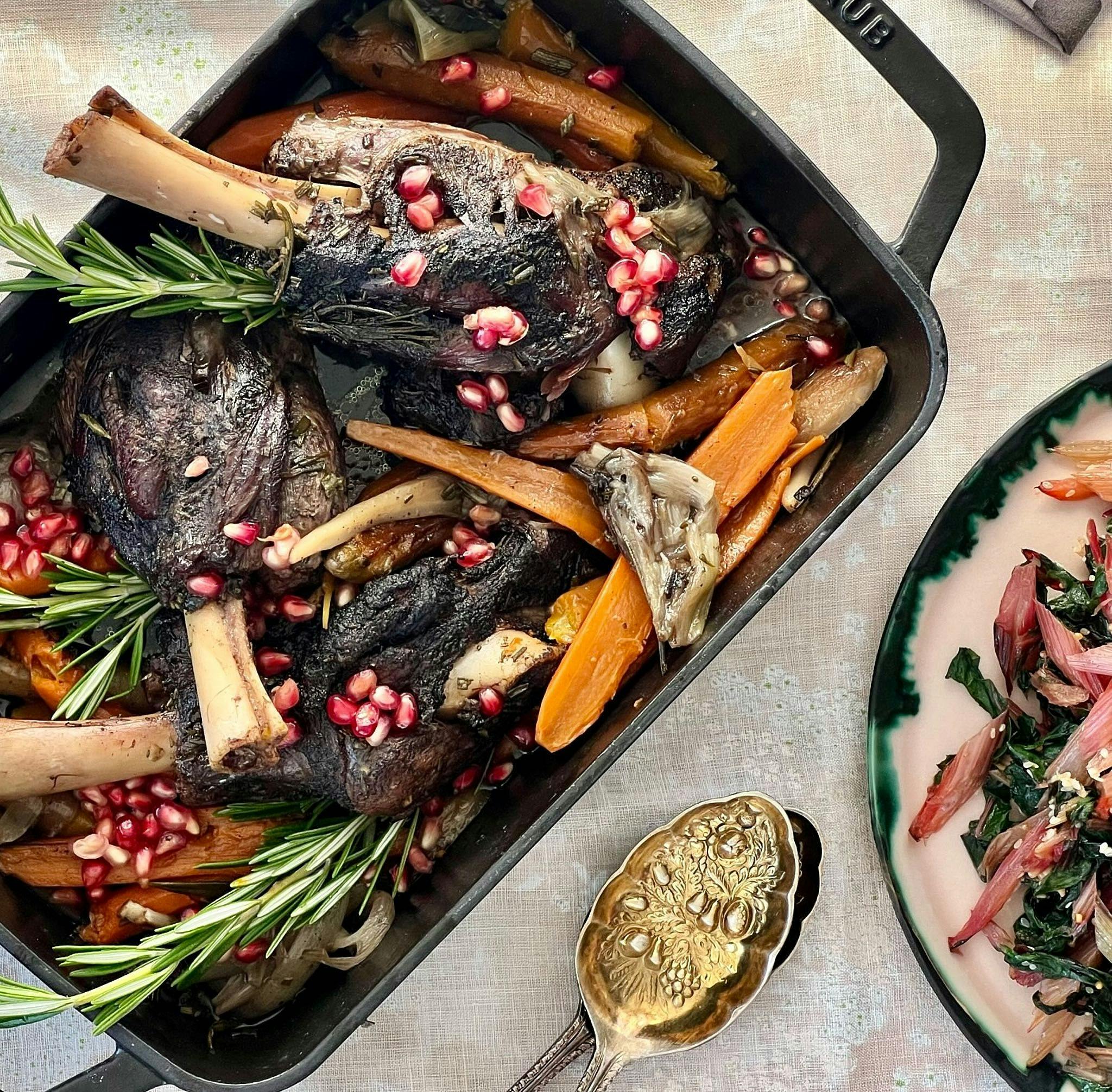 Braised lamb in serving dish with pomegranate seeds, carrots, and rosemary